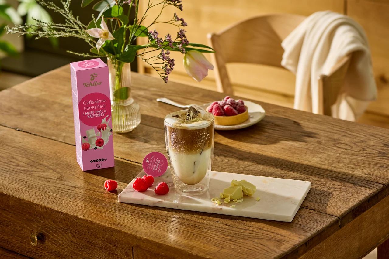 Sur une table en bois, un verre de café, des morceaux de chocolat blanc et des framboises sont posés sur une planche de marbre. À l'arrière-plan, on peut voir un paquet de Cafissimo Flavoured Espresso – White Choc & Raspberry – 10 capsules.