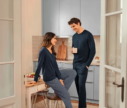 Dans une cuisine, une femme assise sur une table portant un pyjama bleu marine rayé et un haut bleu marine et un homme tenant une tasse et portant un pyjama bleu marine.
