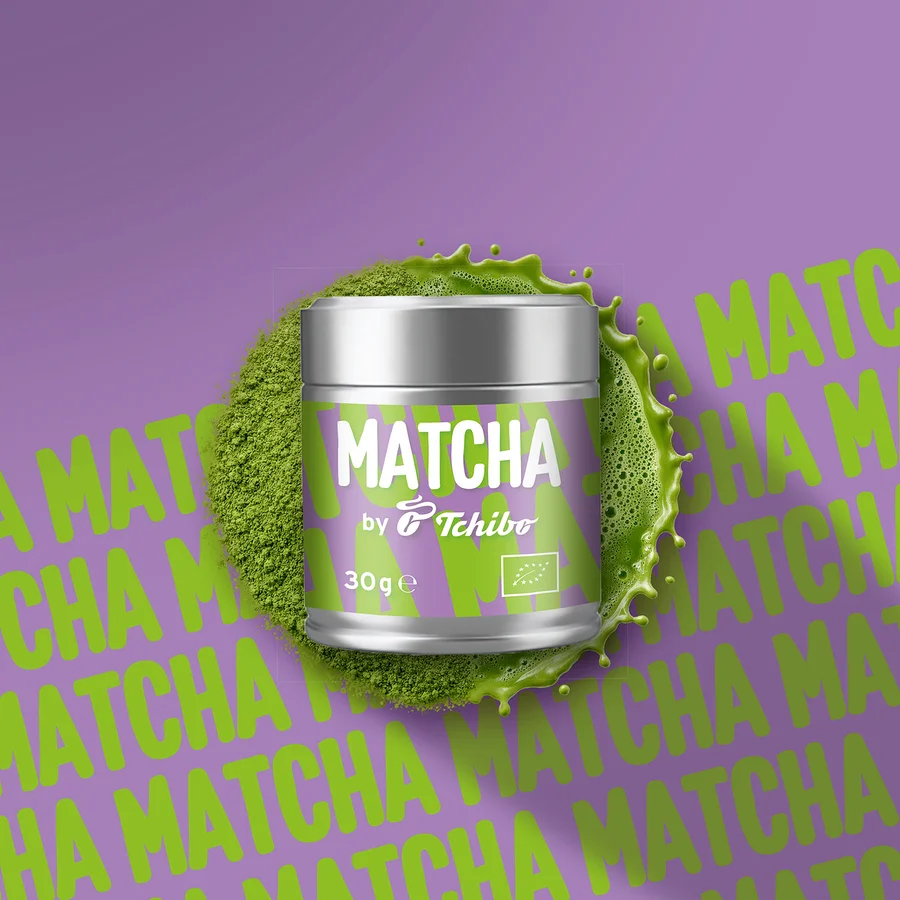 Boîte de Matcha Tchibo argentée avec étiquette violette, entourée de poudre de matcha verte et d'éclaboussures, sur fond violet avec texte Matcha répété.