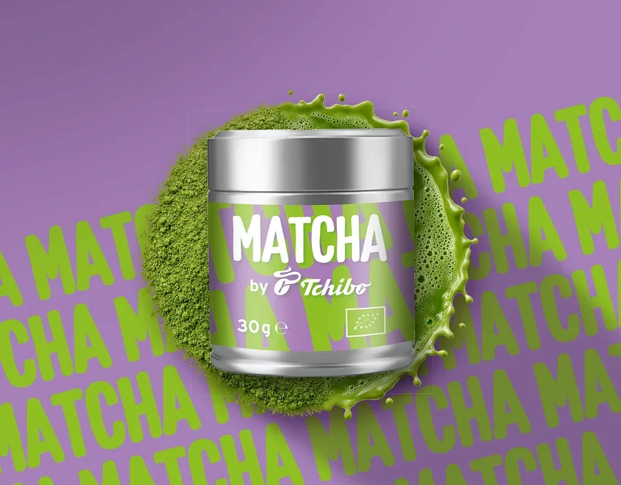 Boîte de Matcha Tchibo argentée avec étiquette violette, entourée de poudre de matcha verte et d'éclaboussures, sur fond violet avec texte Matcha répété.