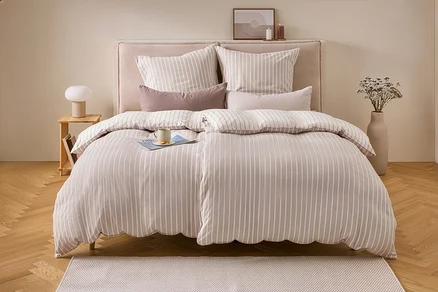 Un lit avec une parure de lit en flanelle rayée beige et blanche, taille double, avec un magazine et une tasse. À gauche, une table de chevet avec une lampe, à droite, un vase avec des fleurs séchées.
