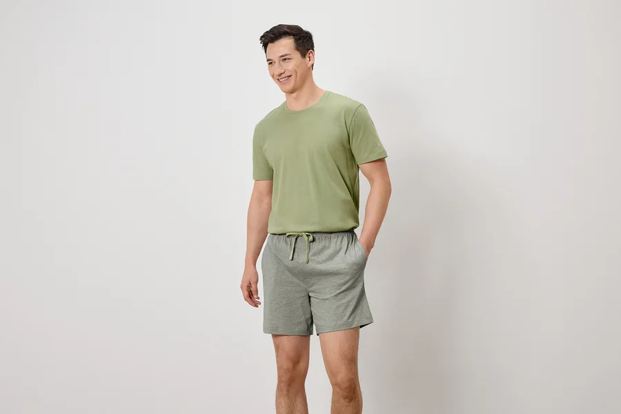 Homme souriant portant un t-shirt vert olive et un short gris avec cordon de serrage, les mains dans les poches.