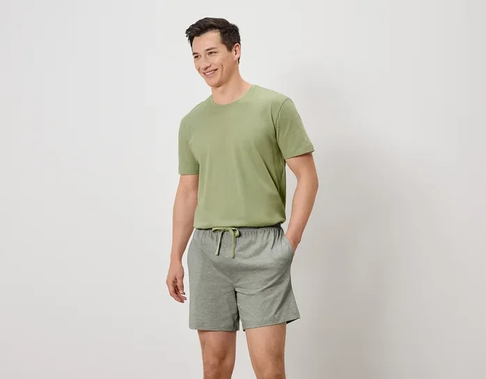 Homme souriant portant un t-shirt vert olive et un short gris avec cordon de serrage, les mains dans les poches.