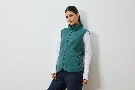 Femme avec queue de cheval portant un gilet en polaire vert sur un t-shirt blanc à manches longues et un pantalon bleu marine.