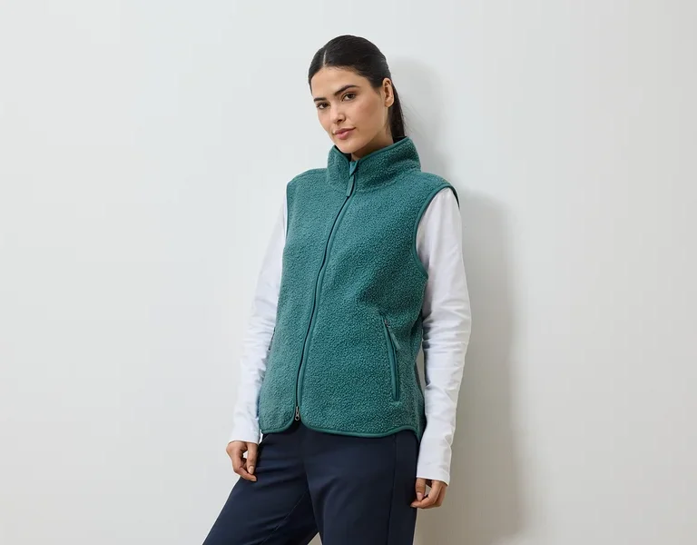 Femme avec queue de cheval portant un gilet en polaire vert sur un t-shirt blanc à manches longues et un pantalon bleu marine.