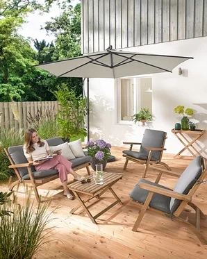 Une femme est assise sur un ensemble salon de jardin pliable avec coussins premium, elle lit et est entourée d'une tablette de jardin et de 2 coussins d’extérieur avec texture lin.