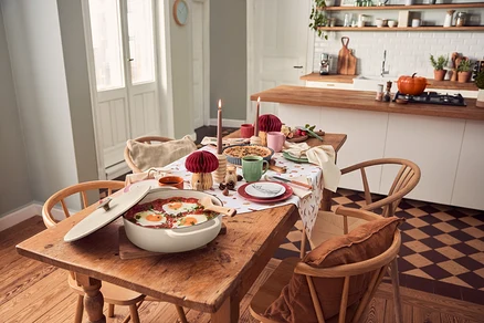 Une cocotte basse en fonte crème avec des œufs au plat dans une sauce rouge est posée sur une table en bois dressée avec des bougies et de la vaisselle. Une cuisine est visible en arrière-plan.
