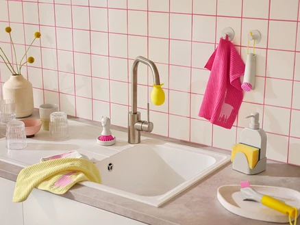 Un évier de cuisine avec un mur carrelé blanc et des joints roses. Sur le comptoir se trouvent des torchons jaunes et roses à motif de lama, des brosses à vaisselle et un distributeur de savon avec porte-éponge.