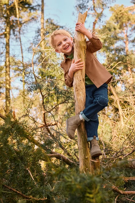Un jeune garçon souriant grimpe à un tronc d'arbre dans une forêt, portant une veste marron, un t-shirt vert, un jean bleu avec des patchs aux genoux et des bottes marron clair.