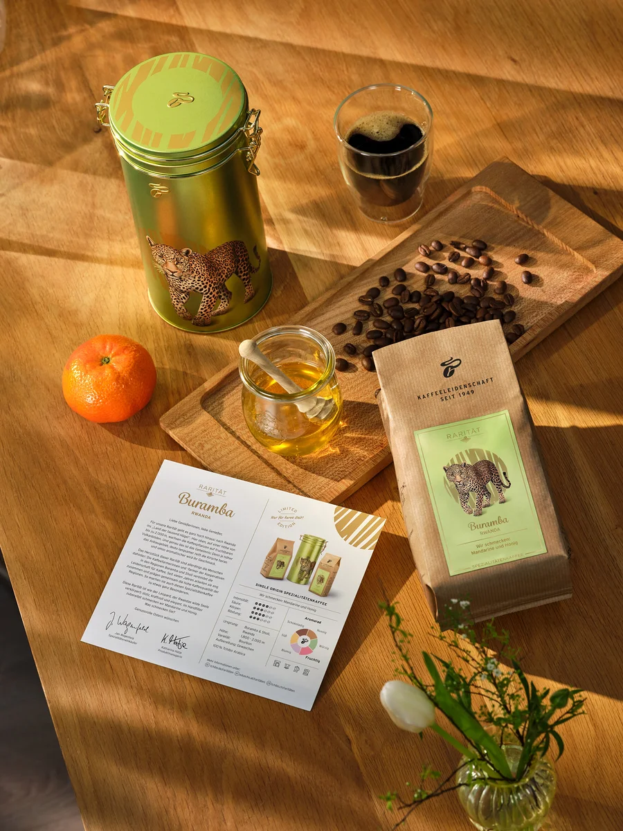 Sur une table en bois se trouvent une boîte, un paquet et un papier de café, une tasse de café, une mandarine, du miel et un vase de tulipes.