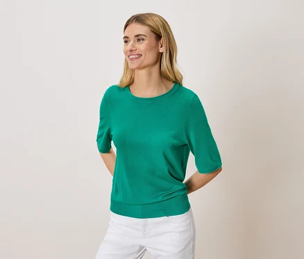 Femme blonde portant un pull vert et un pantalon blanc.