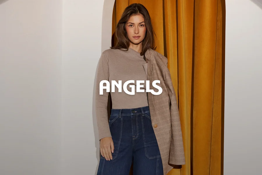 Une femme vêtue d'un haut beige à manches longues et d'un jean bleu foncé, avec un blazer à carreaux marron drapé sur son épaule, se tient devant un mur blanc et un rideau doré. Le mot ANGELS est superposé.