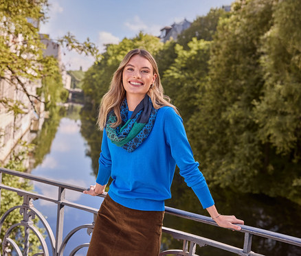 Femme souriante en haut bleu, jupe marron et écharpe à motifs, portant des Créoles doubles en argent 925 rhodié, appuyée sur un pont au-dessus d'une rivière.