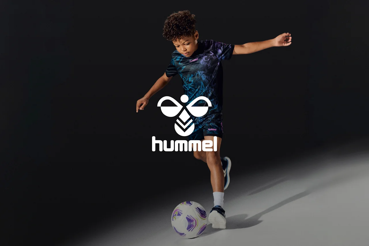 Jeune garçon en tenue de sport à motifs noirs et violets, frappant un ballon de football blanc et violet. Un logo Hummel blanc est superposé.