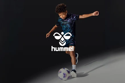 Jeune garçon en t-shirt et short sombres à motifs, frappant un ballon de football blanc et violet, avec le logo blanc Hummel.