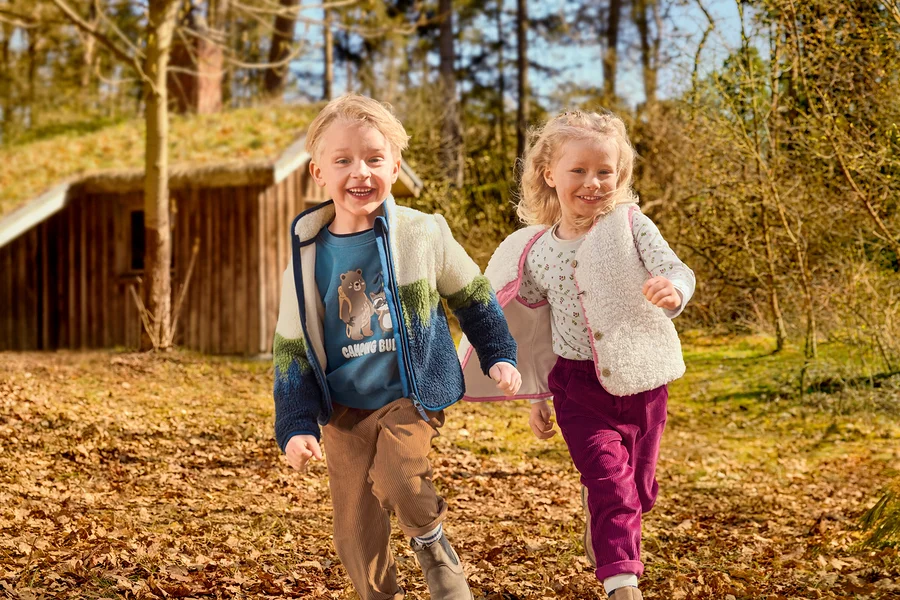 Deux enfants souriants, un garçon et une fille, courent dans un espace extérieur couvert de feuilles. Le garçon porte un t-shirt bleu à motif, une veste polaire colorée et un pantalon en velours côtelé marron. La fille porte un t-shirt à motifs, un gilet moelleux crème et un pantalon en velours côtelé magenta. Une maison en bois avec un toit en herbe est en arrière-plan.
