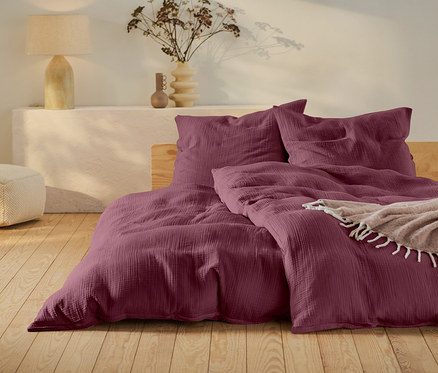 Linge de lit bordeaux sur un lit avec cadre en bois, un pouf beige et une lampe sur une table blanche.