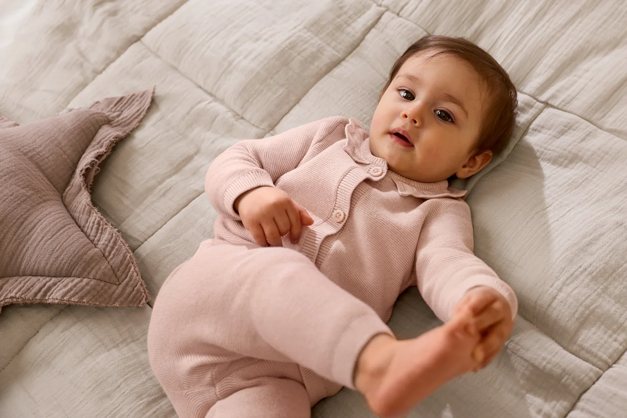 Un bébé vêtu d'un cardigan en tricot rose et d'un pantalon assorti est allongé sur une couverture texturée gris clair, regardant vers le haut. Un coussin rose en forme d'étoile est à côté du bébé.