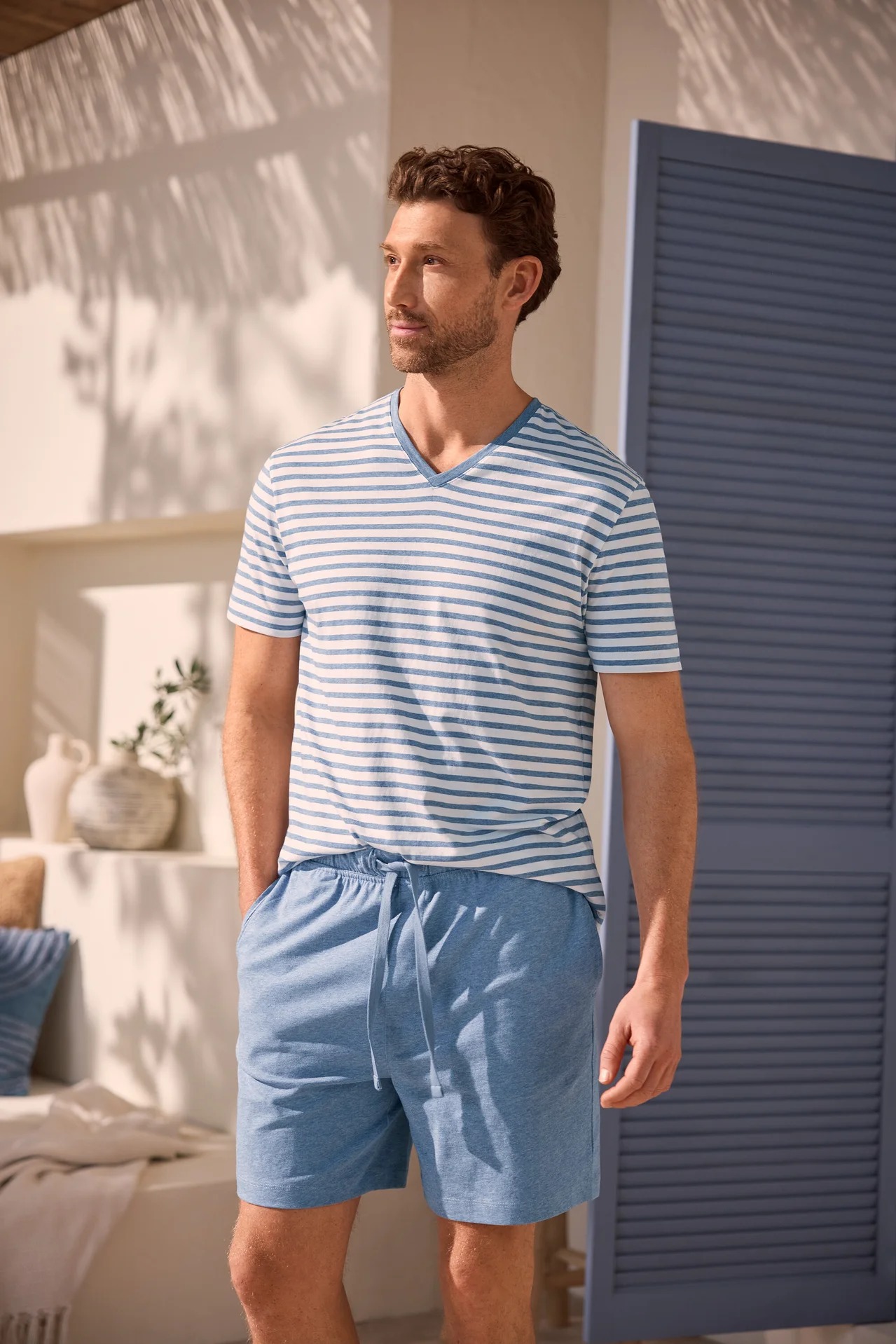 Un homme aux cheveux bruns bouclés et à la barbe porte un t-shirt rayé bleu et blanc à col en V et un short bleu clair. Il se tient les mains dans les poches devant un mur blanc ombragé et une porte bleue.