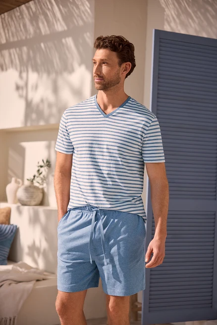 Un homme aux cheveux bruns bouclés et à la barbe porte un t-shirt rayé bleu et blanc à col en V et un short bleu clair. Il se tient les mains dans les poches devant un mur blanc ombragé et une porte bleue.
