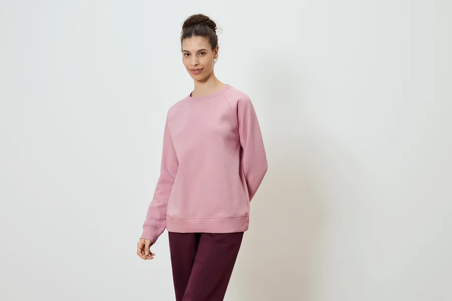 Une femme portant un sweat-shirt rose et un pantalon bordeaux sur un fond blanc.
