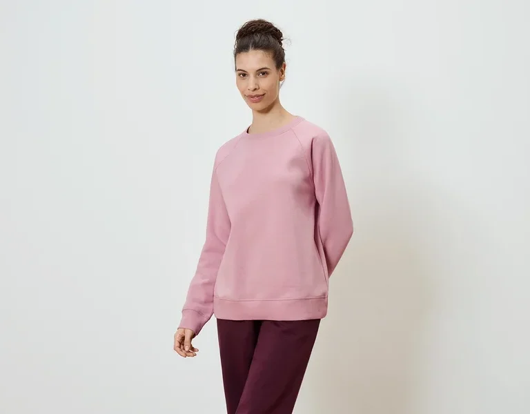 Une femme portant un sweat-shirt rose et un pantalon bordeaux sur un fond blanc.