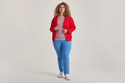 Une femme aux cheveux bouclés se tient debout, vêtue d'un cardigan rouge déboutonné sur un t-shirt rayé, d'un pantalon bleu clair et de baskets blanches.