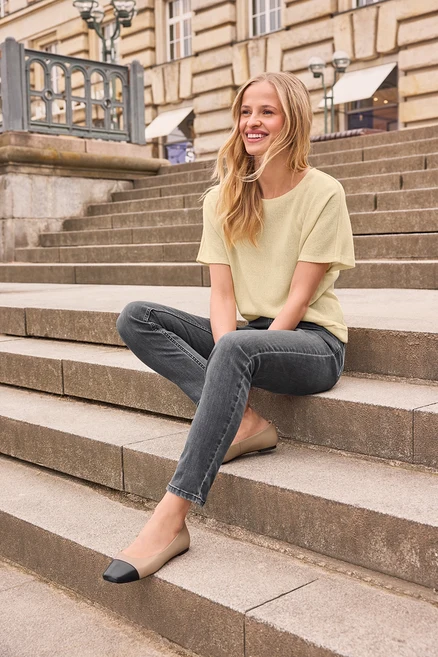 Une jeune femme blonde souriante est assise sur des marches en pierre, vêtue d'un haut jaune clair, d'un jean gris foncé et de ballerines en cuir beiges et noires.