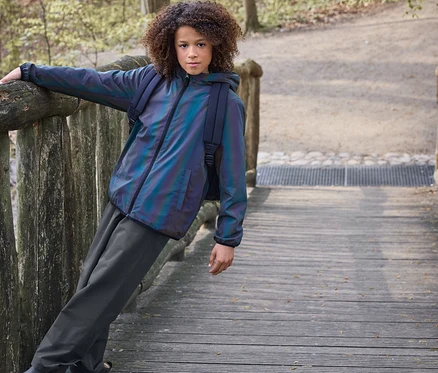 Un enfant aux cheveux bouclés s'appuie sur une balustrade en bois sur un pont. Il porte une veste irisée, un pantalon foncé et des bottines Chelsea de pluie pour enfant, doublées, marron.