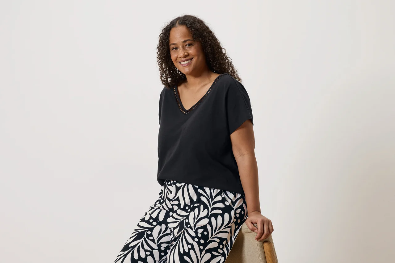 Une femme souriante aux cheveux bouclés, portant un t-shirt noir à col en V et un pantalon à motifs, assise sur une chaise.