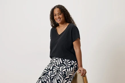 Une femme souriante aux cheveux bouclés, portant un t-shirt noir à col en V et un pantalon à motifs, assise sur une chaise.