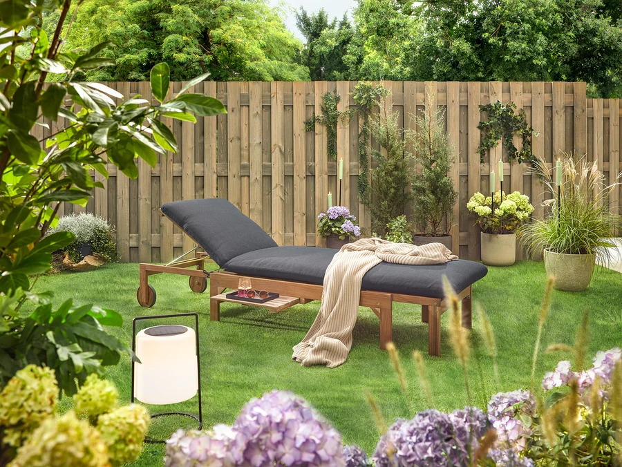 Scène de jardin avec une chaise longue en bois avec un rembourrage noir, une couverture, une boisson, des lunettes de soleil et une lanterne sur une pelouse devant une clôture en bois.
