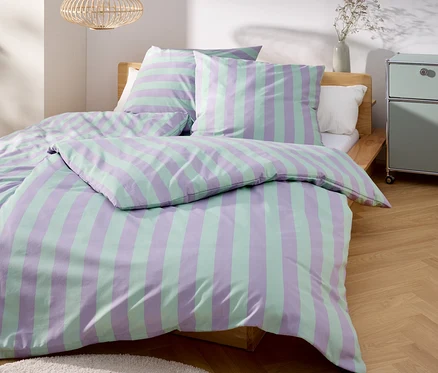 Un lit avec des draps rayés violets et verts se trouve dans une pièce lumineuse.