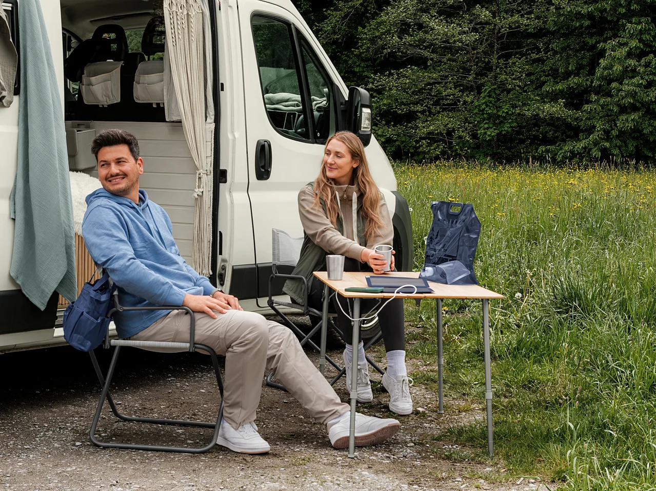 Un homme en sweat-shirt à capuche bleu clair et une femme en veste en sweat beige et gilet hybride kaki sont assis devant un camping-car. Un gilet pliable bleu foncé est posé sur une table.
