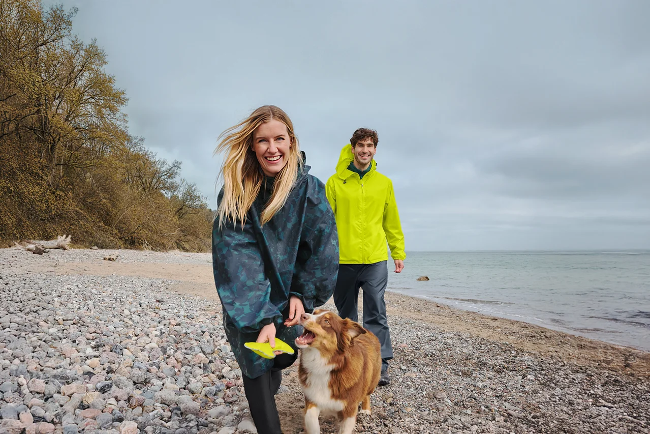 Une femme souriante en poncho de pluie foncé tient un frisbee jaune en marchant avec un chien marron et blanc sur une plage de galets. Un homme souriant en veste de pluie jaune marche derrière eux, avec la mer et un ciel nuageux en arrière-plan.