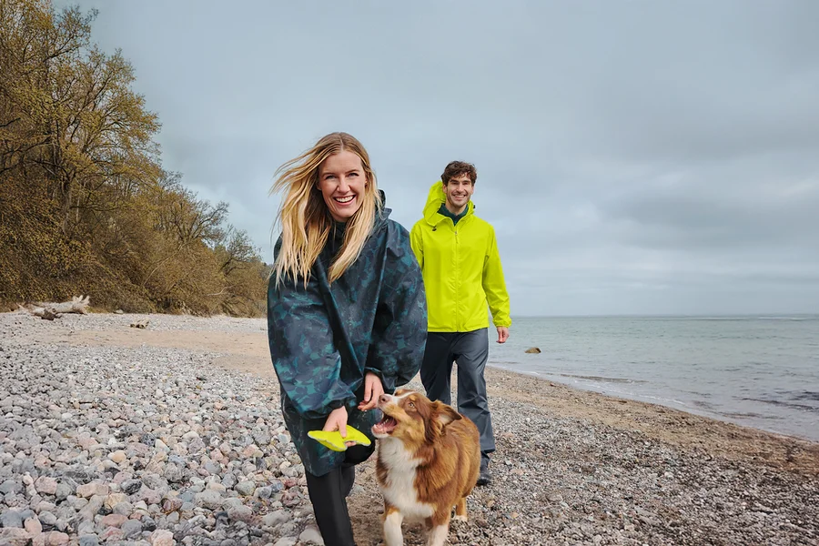Une femme souriante en poncho de pluie foncé tient un frisbee jaune en marchant avec un chien marron et blanc sur une plage de galets. Un homme souriant en veste de pluie jaune marche derrière eux, avec la mer et un ciel nuageux en arrière-plan.