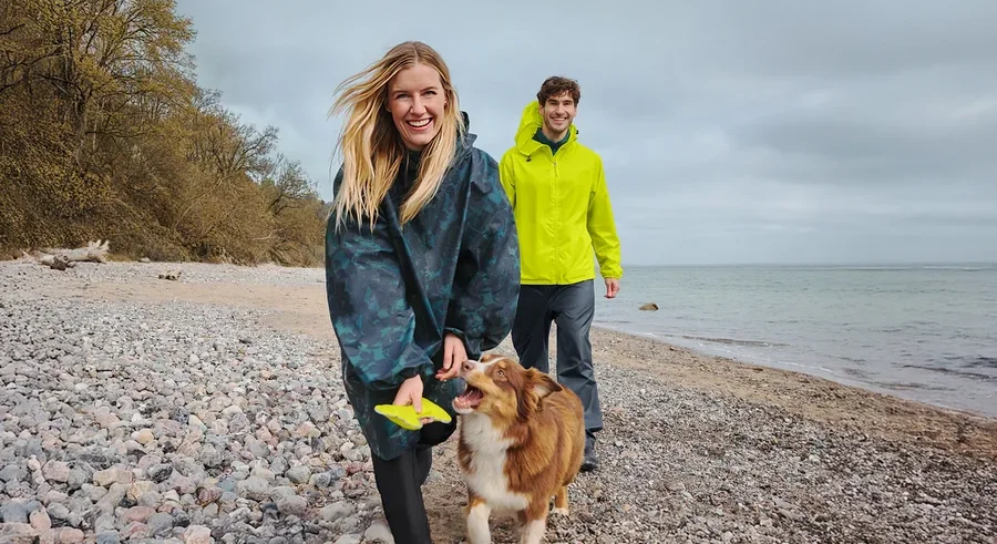 Une femme souriante en poncho de pluie foncé tient un frisbee jaune en marchant avec un chien marron et blanc sur une plage de galets. Un homme souriant en veste de pluie jaune marche derrière eux, avec la mer et un ciel nuageux en arrière-plan.