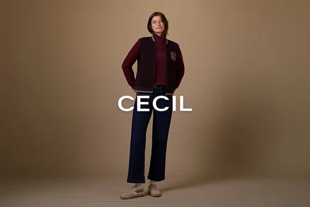 Une femme se tient les mains dans les poches, vêtue d'un col roulé rouge foncé, d'un blouson aviateur marron foncé avec un écusson blanc en forme de « B », d'un jean évasé bleu foncé et de baskets rose clair avec des chaussettes blanches, sur un fond marron uni.