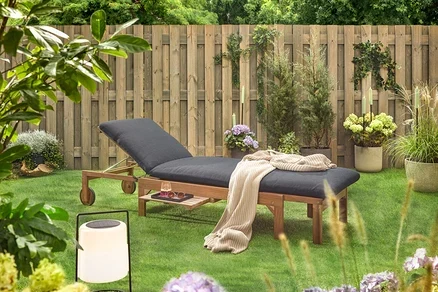 Scène de jardin avec une chaise longue en bois avec un rembourrage noir, une couverture, une boisson, des lunettes de soleil et une lanterne sur une pelouse devant une clôture en bois.
