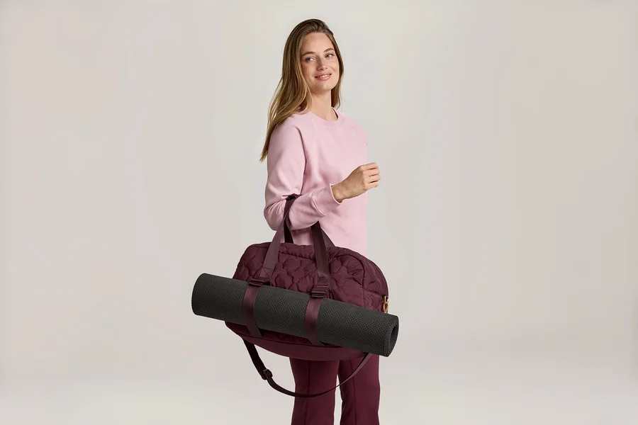 Une femme portant un pull rose et un pantalon bordeaux porte un sac de sport bordeaux avec un tapis de yoga noir sur l'épaule.