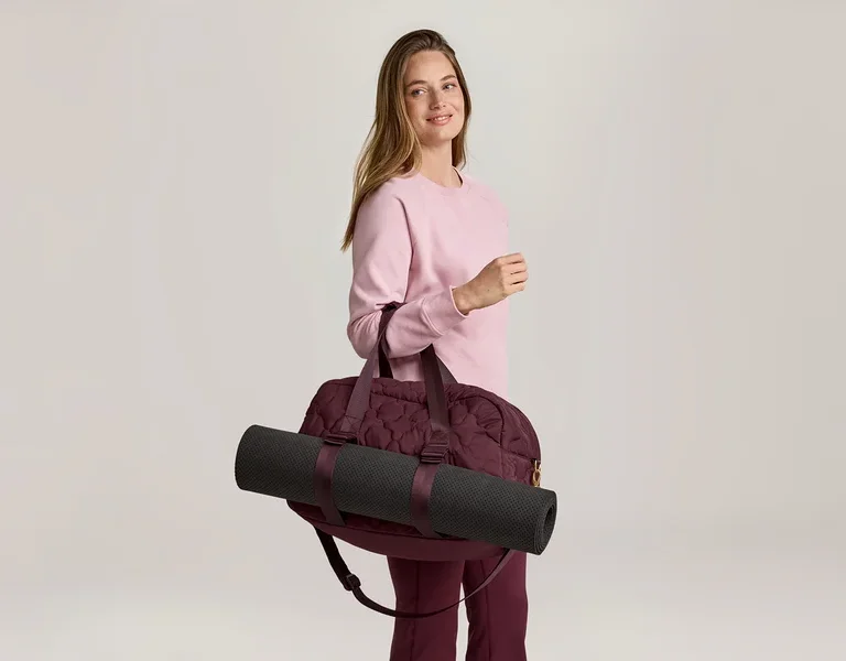 Une femme portant un pull rose et un pantalon bordeaux porte un sac de sport bordeaux avec un tapis de yoga noir sur l'épaule.