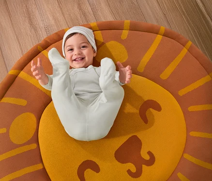 Un bébé souriant est allongé sur un tapis de chambre d’enfant « Lion » et porte un ensemble pour bébé 3 pièces.