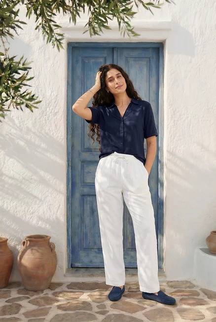 Une femme pose devant une porte bleue, portant un chemisier bleu à boutons, un pantalon en lin blanc, des mocassins en daim, des clous d'oreilles et un bracelet de tennis.