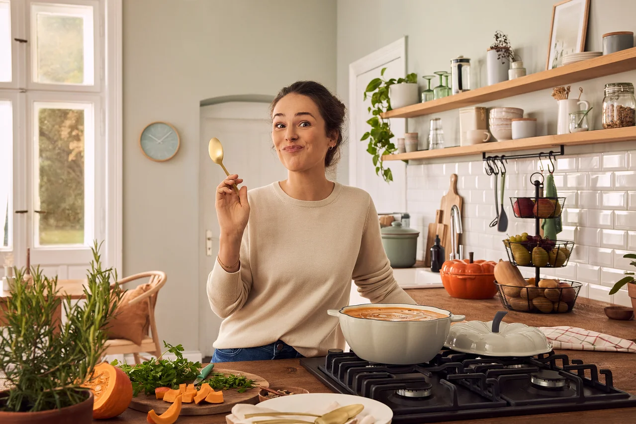Une femme souriante en pull crème tient une cuillère dorée dans une cuisine. Sur le comptoir en bois, un plat en fonte en forme de courge crème est sur une cuisinière à gaz, à côté d'un plat en fonte en forme de courge orange, de la courge coupée et des herbes sur une planche à découper, ainsi qu'un plateau à étages avec des fruits. Un ensemble de couverts couleur or champagne est posé sur une assiette.