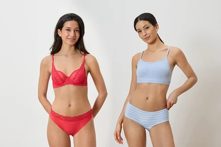 Deux femmes posent côte à côte. La femme de gauche porte un soutien-gorge en dentelle rouge et une culotte rouge assortie. La femme de droite porte une brassière bleu clair et un shorty rayé bleu clair.