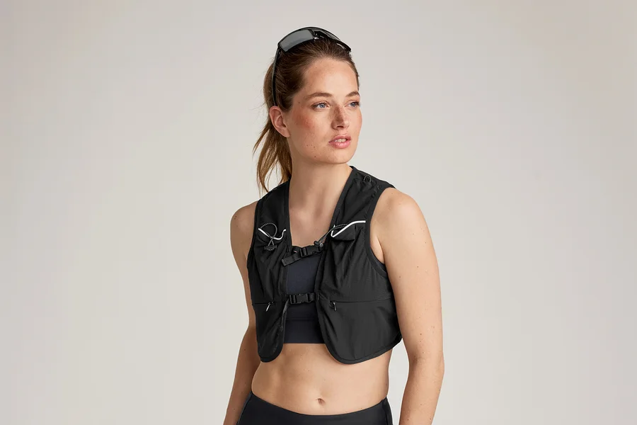 Une femme portant des lunettes de soleil sur la tête porte un gilet de course noir sur un soutien-gorge de sport et un legging.