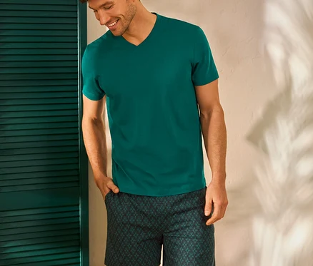 Un homme souriant, la main dans la poche, porte un t-shirt vert et un short à motifs, debout à côté d'un tabouret avec des livres.