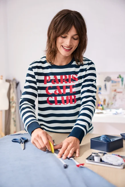 Femme souriante portant un haut rayé bleu marine et blanc avec l'inscription « PARIS CAFE CLUB », marquant un tissu bleu clair avec une craie de tailleur et un mètre sur une table en bois.