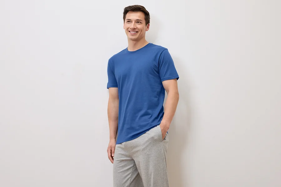Homme souriant en t-shirt bleu et pantalon de survêtement gris posant sur fond blanc.