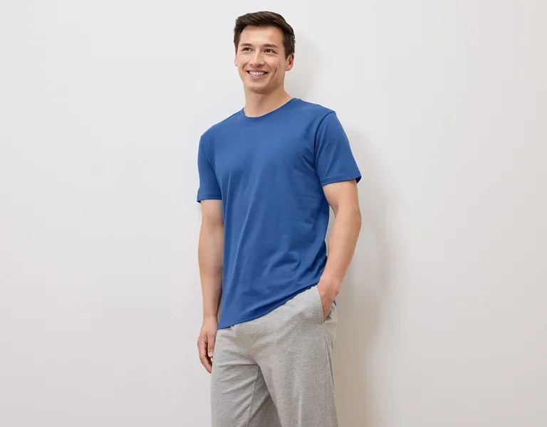 Homme souriant en t-shirt bleu et pantalon de survêtement gris posant sur fond blanc.
